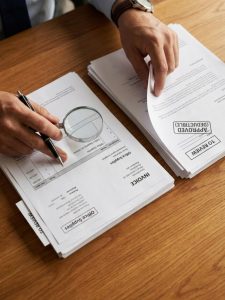 Manos revisando un grupo de facturas y documentos con una lupa y sello de aprobación.
