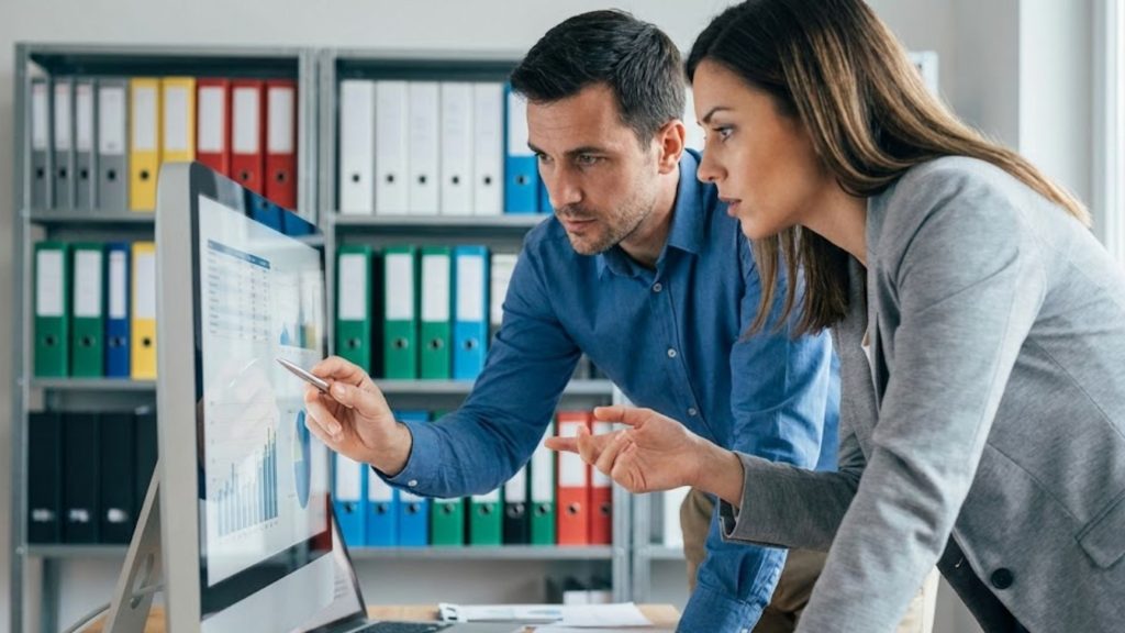 Dos colegas revisan gráficos de datos en una pantalla de ordenador, trabajando juntos en la oficina.